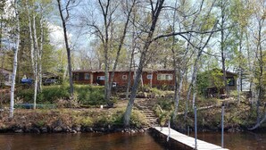 A cabin by a river with a wooden dock and a stone staircase leading to the entrance.