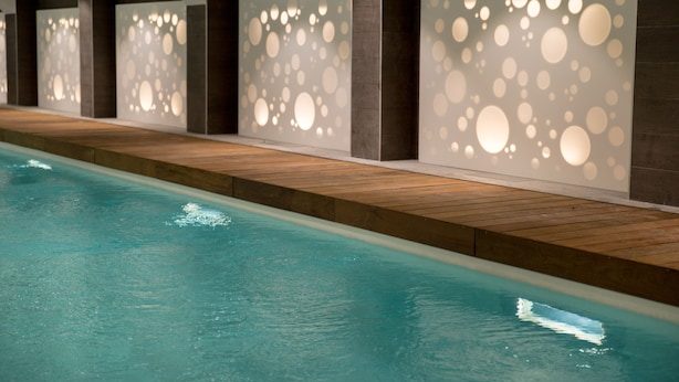 Une piscine dotée d’une terrasse en bois et d’un mur orné de motifs lumineux circulaires.