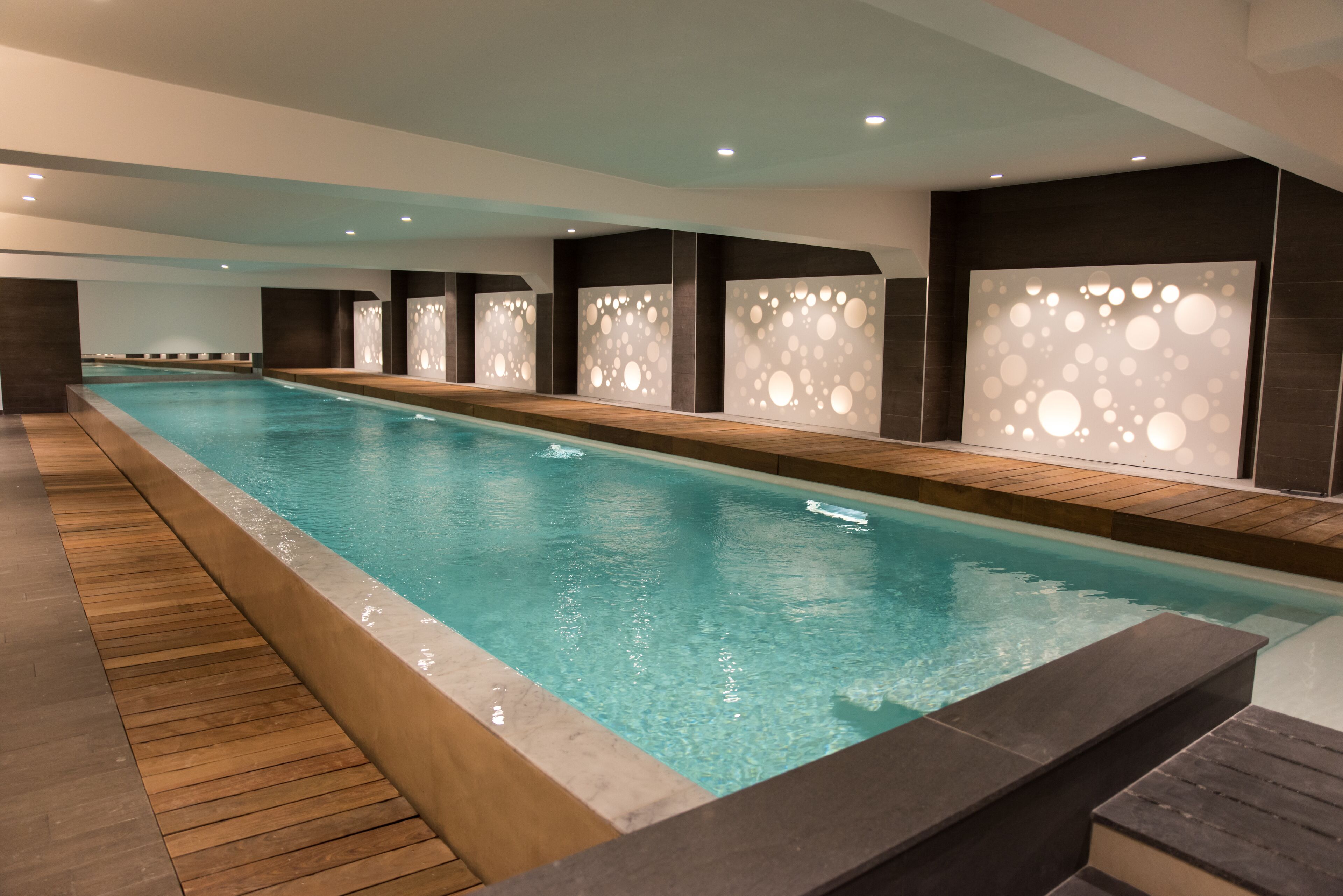 A long indoor swimming pool with a wooden deck and modern wall panels.
