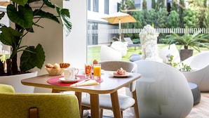 Une table de petit-déjeuner avec des croissants, une boisson aux fruits et un croissant sur une assiette, le tout installé dans un espace extérieur lumineux, avec une clôture blanche et de la verdure en arrière-plan.