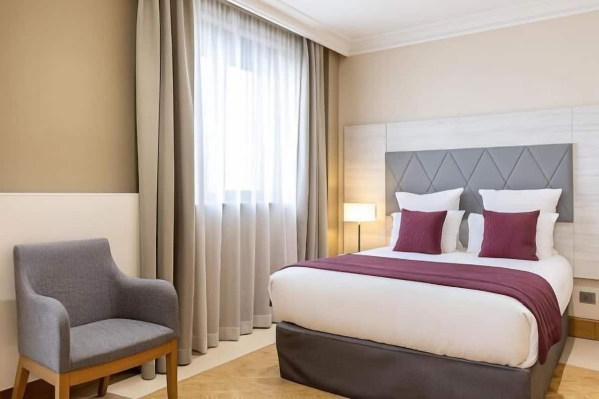 Ein Hotelzimmer mit Bett, Stuhl, Vorhängen und Kopfteil.