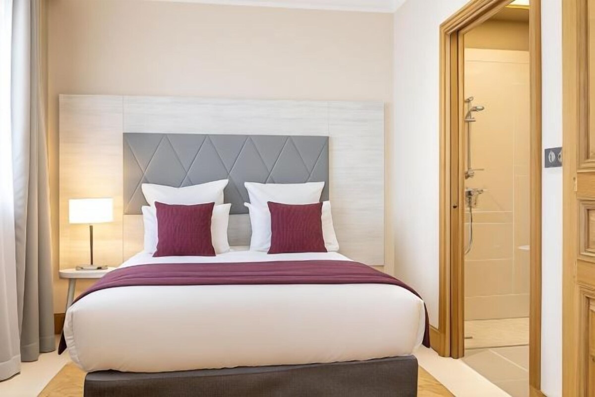 Een hotelkamer met een groot bed, een nachtkastje, een lamp en een houten deur naar een badkamer.