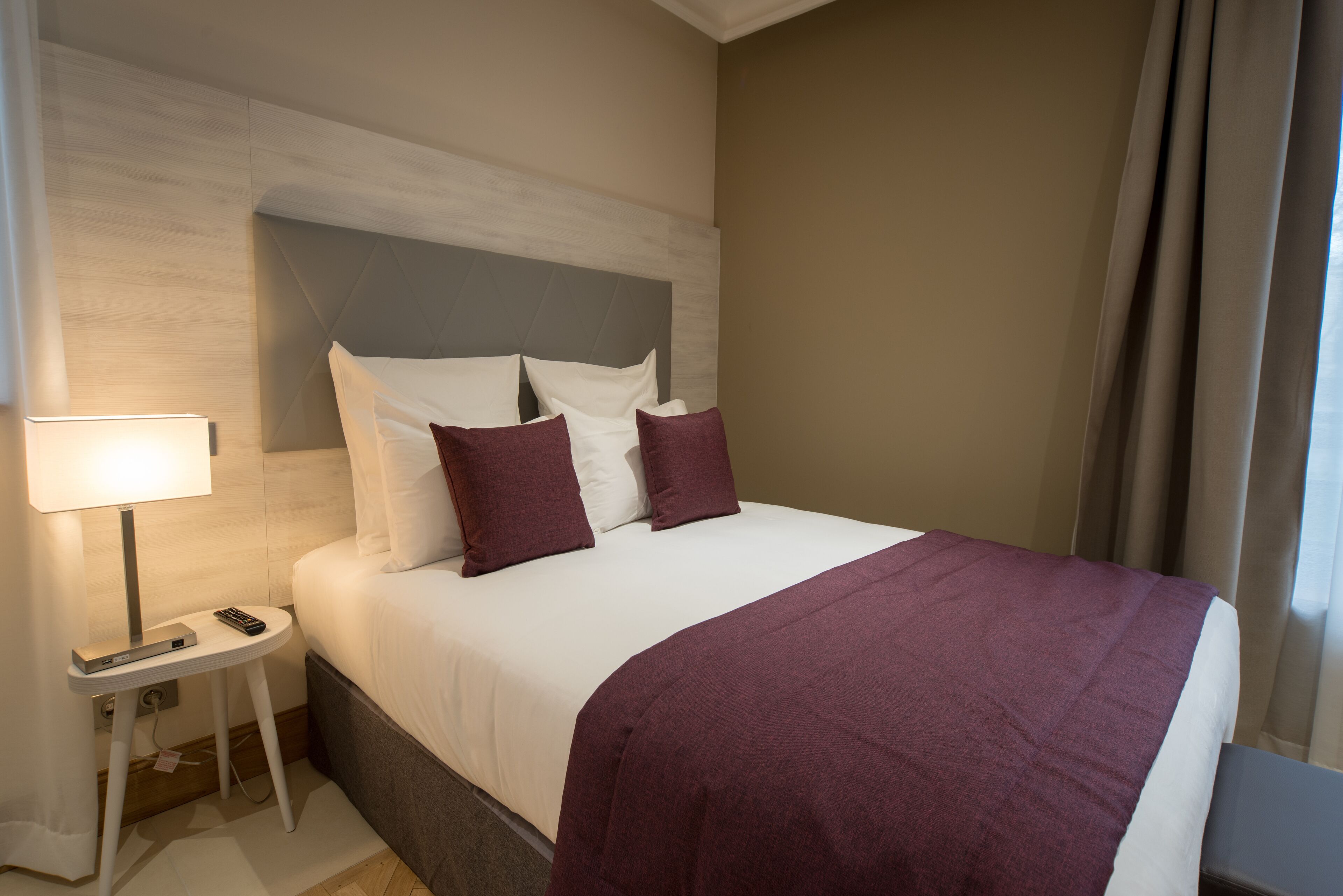Un lit bien fait, recouvert d’une couvre-lit blanche et violette, une table de chevet avec une lampe et une tête de lit moderne.