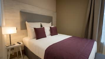 Un lit bien fait, recouvert d’une couvre-lit blanche et violette, une table de chevet avec une lampe et une tête de lit moderne.