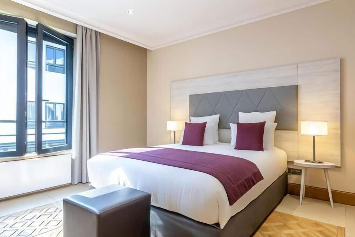 Ein Hotelzimmer mit einem großen Bett, zwei Nachttischlampen, Blick durch ein Fenster und einem kleinen Tisch.