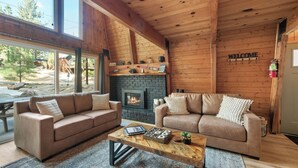 A cozy cabin interior with two sofas facing a fireplace, a coffee table, and a large window overlooking a forested area.