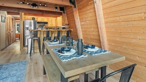 A wooden interior with a dining table set for four, a kitchen in the background, and a sloped ceiling.