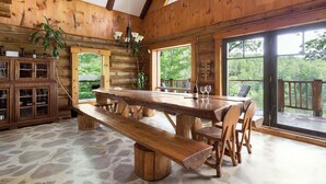 L’intérieur d’un chalet rustique en bois, avec un sol en pierre, une table à manger en bois, des bancs et de grandes fenêtres donnant sur la forêt.