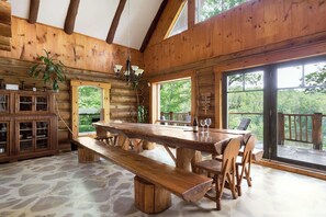 Interior rústico de una cabaña de madera con piso de piedra, mesa de comedor de madera, bancos y ventanas grandes que ofrecen vistas al bosque.