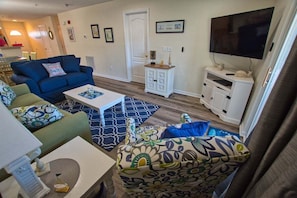 A living room with a blue sofa, a patterned sofa, a white coffee table, a TV on a stand, and a door in the background.