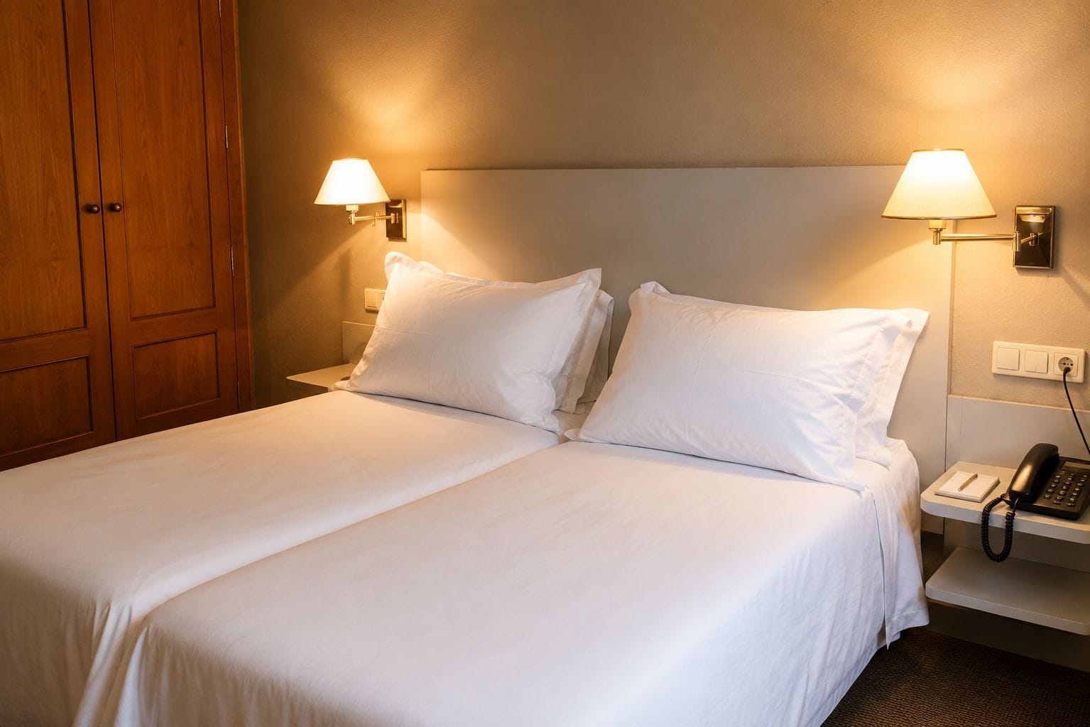 Una habitación de hotel con una cama doble, dos lámparas de noche, un teléfono en la mesita y un armario de madera.
