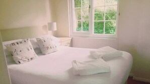 A neatly made bed with white linens and pillows, a bedside lamp, and a window with a view of greenery.