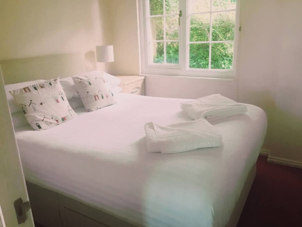 A neatly made bed with white linens and pillows, a bedside lamp, and a window with a view of greenery.