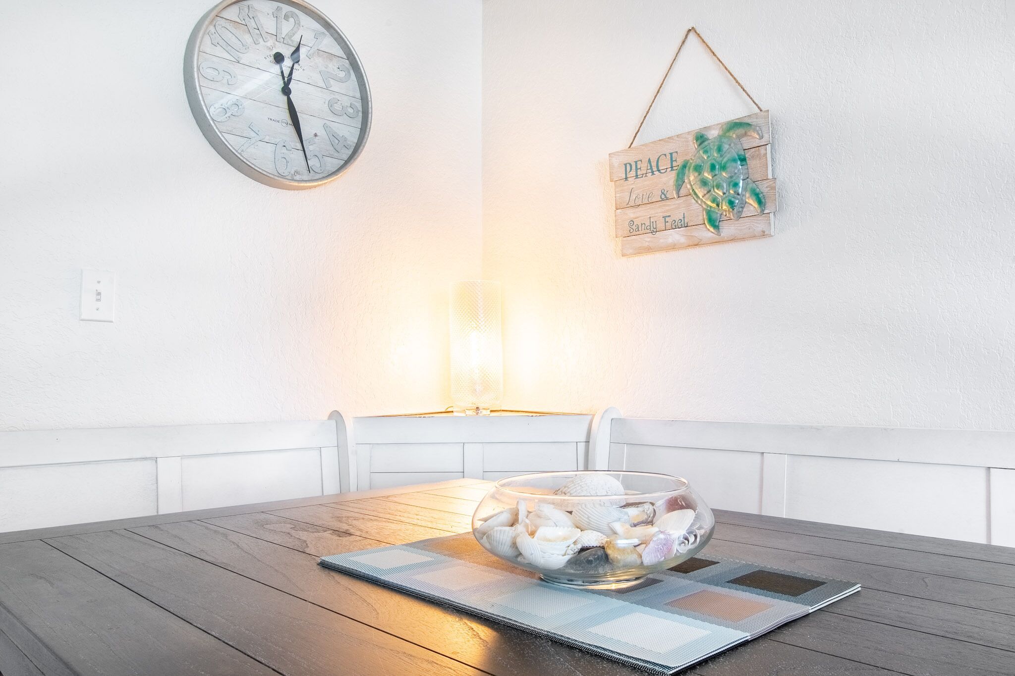 Ein Essbereich mit einem Holztisch, einer Schale Muscheln, einer Wanduhr und einem hängenden Schild mit dem Schriftzug 'PEACE Love & Sandy Feel'.
