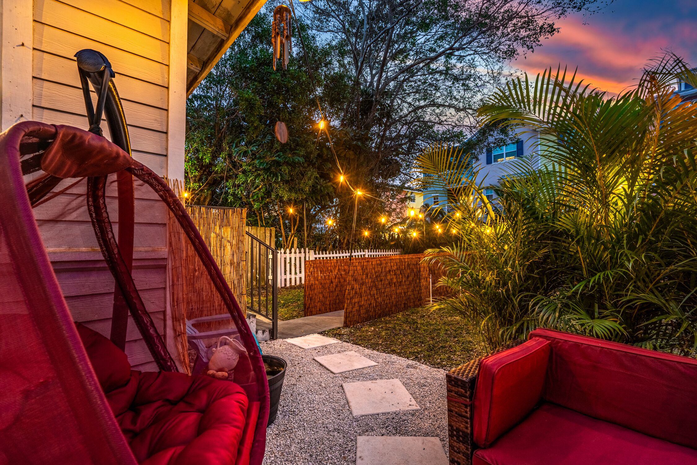 A cozy outdoor seating area with a hammock, a red sofa, and a pathway leading to a wooden fence.