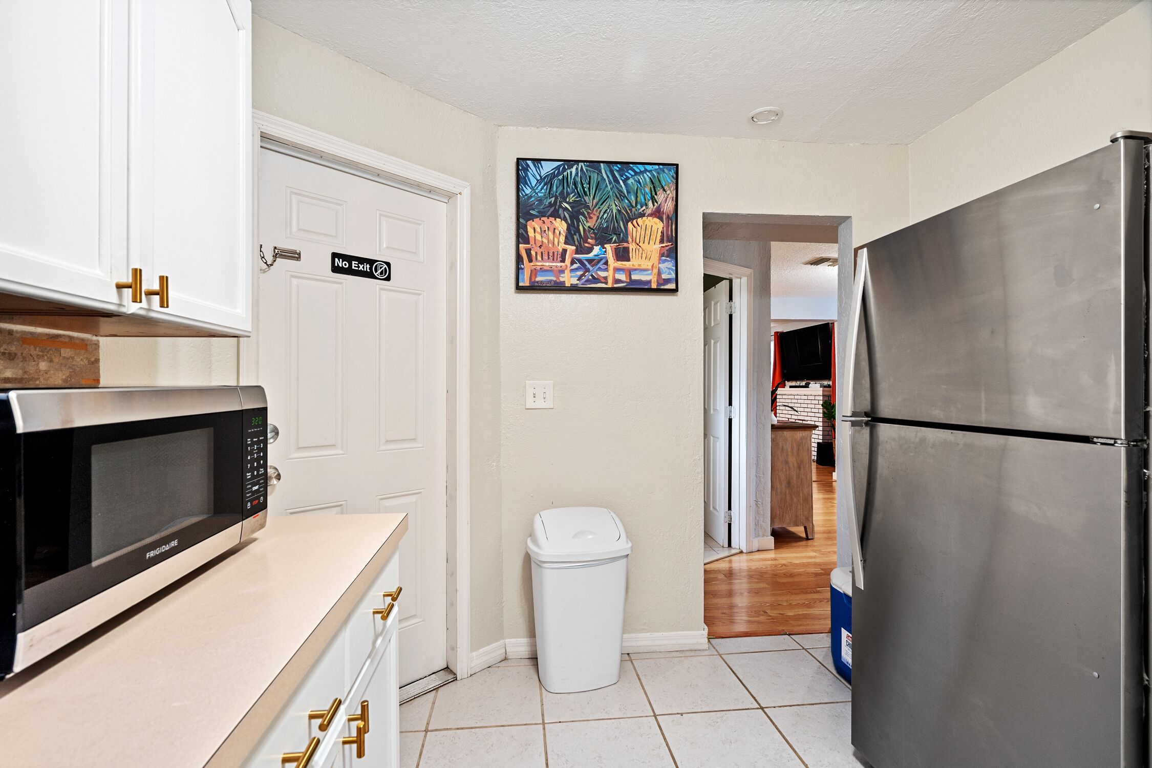 A kitchen with white cabinets, a stainless steel refrigerator, a trash can, and a painting of chairs on a table.