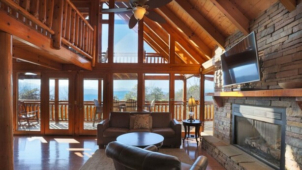 A spacious living area with a stone fireplace, a television mounted on the wall, and a balcony with a view of the mountains. - Secluded Cabin w/Mtn. Views, 3 Decks & Theater (Sevierville)