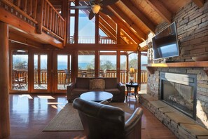 A spacious living area with a stone fireplace, a television mounted on the wall, and a balcony with a view of the mountains. - Secluded Cabin w/Mtn. Views, 3 Decks & Theater (Sevierville)