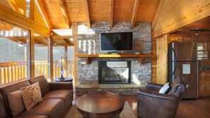 A cozy cabin interior with a stone fireplace, wooden ceiling beams, a television mounted above it, and a refrigerator in the kitchen area. - Secluded Cabin w/Mtn. Views, 3 Decks & Theater (Sevierville)