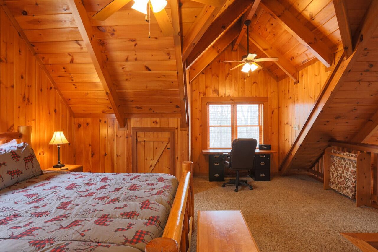 A cozy attic bedroom with a bed, desk, and chair, wooden walls, and a sloped ceiling.