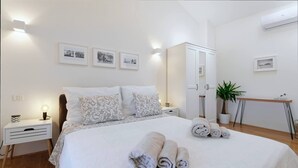 Una cama bien hecha con toallas enrolladas, una lámpara de noche, una mesita con una planta y un armario blanco.