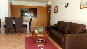 A living room with a brown leather sofa, a dining area with a table and chairs, and a kitchen visible through an open door. - Clube Alvor Ria -Luxury 2 bedroom apartment with stunning views over the harbour (Alvor)