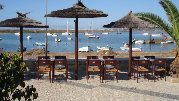 A seaside terrace with thatched umbrellas and wooden chairs overlooking a marina with boats. - Clube Alvor Ria -Luxury 2 bedroom apartment with stunning views over the harbour (Alvor)
