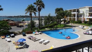 A pool area with lounge chairs and umbrellas, surrounded by palm trees and a view of the sea. - Clube Alvor Ria -Luxury 2 bedroom apartment with stunning views over the harbour (Alvor)