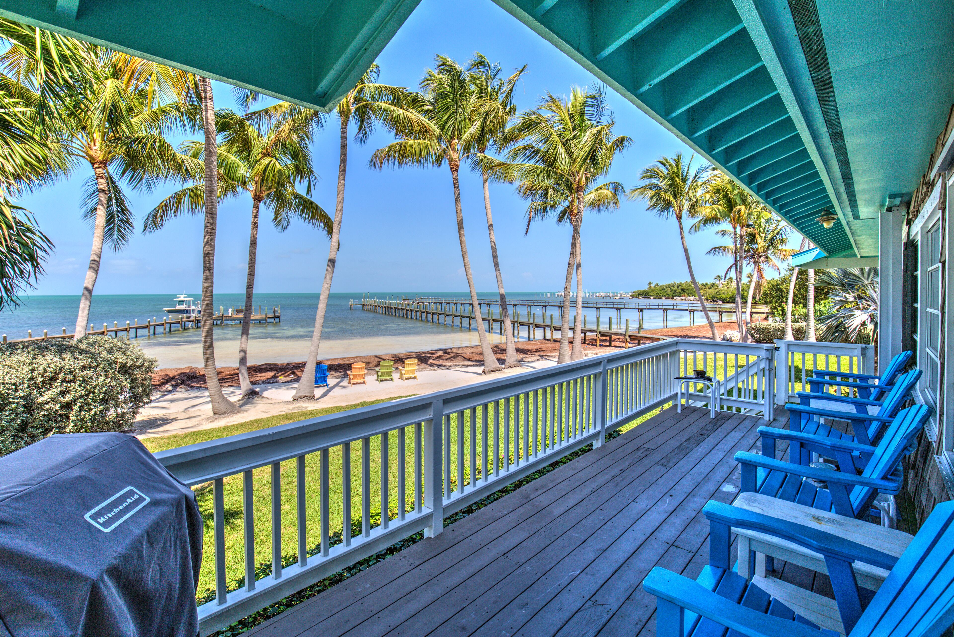 A wooden deck with blue chairs overlooking a beach with palm trees and a pier.