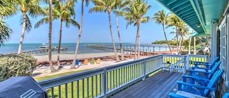 A wooden deck with blue chairs overlooking a beach with palm trees and a pier.