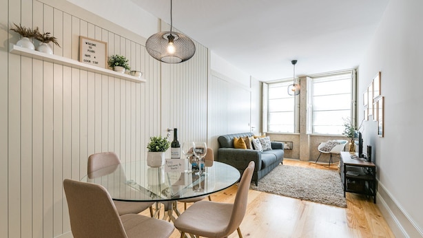 A modern living room with a glass dining table, two sofas, and a coffee table. There are potted plants on shelves and a large window providing natural light. - GuestReady - Oporto Temptation Apartment (Porto)