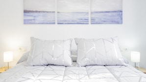 Ein Bett mit einem geometrisch gemusterten Überwurf, zwei weiße Kissen und ein an der Wand hängendes Bild einer Küstenlandschaft.