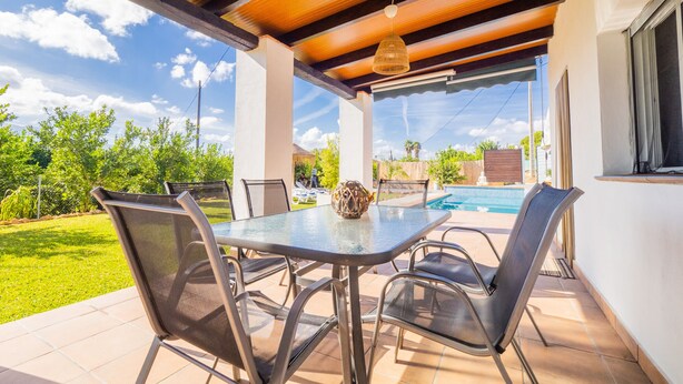 Eine Terrasse im Freien mit einem Glastisch, Metallstühlen, einer Dekovase und Blick auf ein Schwimmbad und Grünflächen.