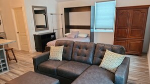 A modern living room with a sectional sofa, ottomans, a bed, a wardrobe, and a small kitchen area. - Track and seasonal rental (Saratoga Springs)
