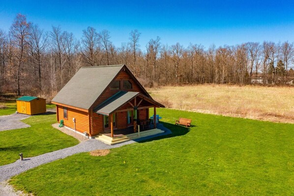 Eine Blockhütte mit Veranda, einem kleinen Schuppen und einem grünen Feld.