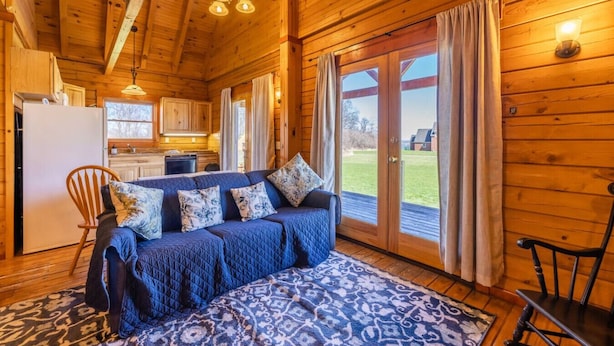 A cozy cabin interior with a blue sofa, wooden walls, a kitchen, and a large sliding glass door to the outdoors.