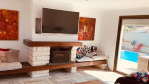 A modern living room with a fireplace, a flat-screen TV mounted above it, and a view of a swimming pool through a glass door. - Luxurious villa in cap d'agde  (Agde)