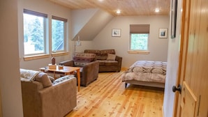 A cozy room with a loft bed, a sofa, and a small table. There are two windows providing natural light and a view of trees. - Aviator Cabin (Talkeetna)