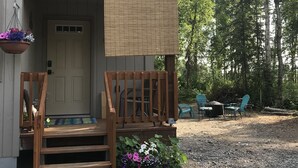 A wooden front porch with steps leading to a door, a hanging flower basket, and a covered area with a woven mat. - Aviator Cabin (Talkeetna)