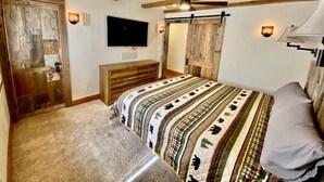 Een slaapkamer met een bed, houten balken in het plafond, een wandtelevisie en een schuifdeur in boerderijstijl.