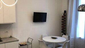 A modern kitchen with a built-in oven, a white dining table with four chairs, a flat-screen TV mounted on the wall, and a bookshelf filled with books. - House Giussano, elegant two-room apartment in the center (Milano)