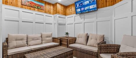 A room with a large flat-screen TV mounted on the wall, wicker furniture, and a wooden paneled ceiling.