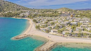 Eine Küstenlandschaft mit einer Reihung von Mobilheimen direkt am Strand, klarem türkisblauem Wasser und einer hügeligen Landschaft im Hintergrund.
