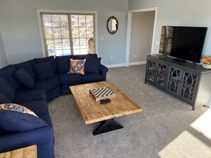 Una sala de estar con un sofá modular azul oscuro, una mesa de centro de madera con un tablero de ajedrez, un televisor sobre un mueble y un ventanal con vista a árboles.