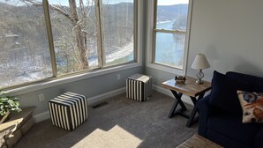 Ein gemütliches Zimmer mit großen Fenstern, einem blauen Sofa, gestreiften Hocker und einem kleinen Holztisch mit einer Lampe.