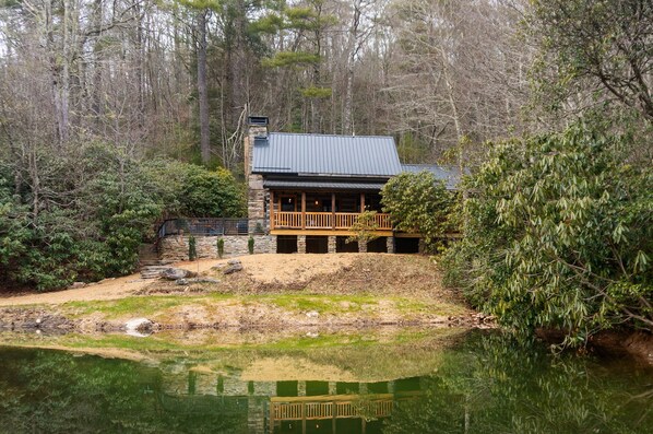 A cabin with a stone foundation, a wooden deck, and a pond in front.