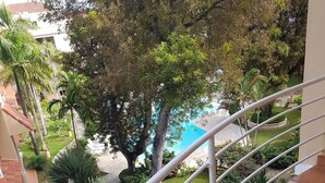 Un balcone con pavimento piastrellato e ringhiera bianca, affacciato su un giardino curato con palme e una piscina.