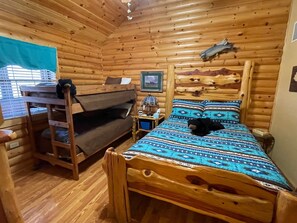 Ein Holzhütten-Zimmer mit einem Etagenbett, einem kleinen Tisch, einem Nachttisch und einem Bett mit gemustertem Überwurf.