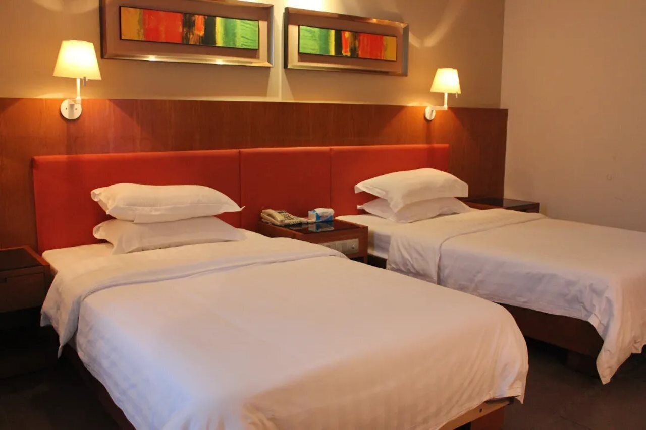 Ein Hotelzimmer mit zwei Einzelbetten, jeweils mit weißen Bettwäsche und Kissen. Es gibt zwei Nachttische, einer mit einer Lampe und der andere mit einem Telefon und einem kleinen Gegenstand. Das Zimmer verfügt über ein Holz-Kopfteil und zwei gerahmte Kunstwerke über den Betten.
