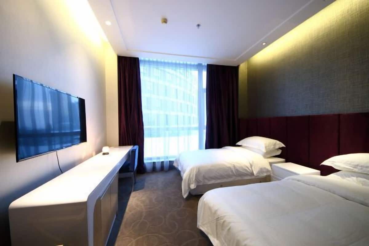 A hotel room with two beds, a desk, a flat-screen TV, and a large window with curtains.